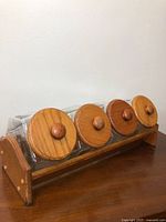 Side angle showing four clear square glass canisters with round wooden lids and knob handles, placed in a wooden holder.