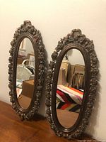 Two oval mirrors with detailed floral plastic frames in bronze finish, placed side-by-side on a wood surface showing front views