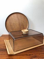 Plastic cheese tray with transparent lid on beige base and round teak serving plate behind it, showing size and materials.