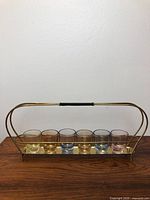 Front view of brass shot glass caddy holding seven colored shot glasses on wooden surface against plain white background.