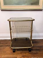 Front view of small brass side table with smoke glass top and bottom shelves, middle brass wire rack shelf, and plastic casters under wooden floor and framed artwork background.