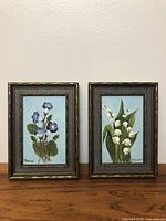 Two small wood framed flower paintings side by side on a wooden surface.