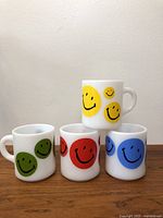 Front view of 4 milk glass mugs with yellow, green, red, and blue smiley face designs arranged on wooden surface