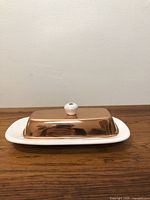 Front view of vintage copper butter dish with ceramic base placed on wooden surface against white wall.