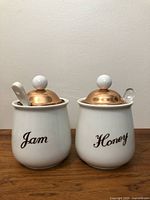 Front view of jam and honey jars showing white ceramic bodies with copper lids and labeled text.