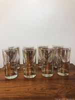 Eight clear highball style cocktail glasses with gold geometric diamond patterns on a wooden surface against a white wall.