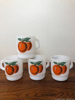 Four vintage milk glass mugs arranged with one on top and three below, showing the orange fruit design on opaque white glass.