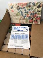 Partial view of latch hook canvas with floral pattern, box containing yarn rolls, and instruction sheet