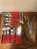 Full layout showing all cutlery pieces with red handles, fondue forks with wooden handles on teak platter.