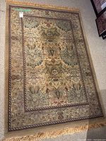 Full view of the rectangular patterned rug with beige background and green, brown, burgundy floral and leaf designs. Fringe visible on shorter sides.