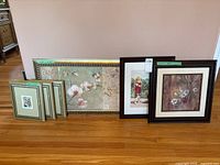 Six framed artworks arranged leaned against a wall on hardwood floor, including the largest floral print and the signed watercolour with a child