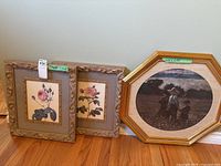 Three framed pictures: two floral prints and one picture of children in a meadow, all in gold frames on wood floor.