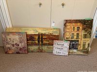 Four canvas art pieces arranged on floor against a door showing different scenes including floral, landscape, building facade, and text art.