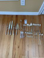 Full assortment of flatware pieces spread on hardwood floor showing knives, forks, various spoons, and serving utensils.