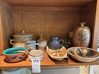 Wooden shelf holding an assortment of pottery including lidded jars, mugs, a vase, a shallow patterned bowl, and a turquoise bowl with bird motifs and orange handle.