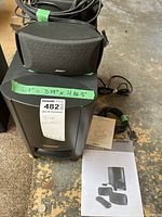 Bose Acoustimass module and satellite speakers with cables and user manual arranged on concrete floor showing size tag and labeled 'Bose Equipment'.