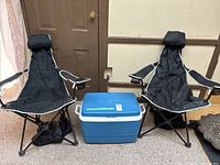 Front view of two black folding lounger chairs with headrests and cup holders, and blue Rubbermaid cooler between them.