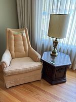 Cream fabric chair, hexagon dark wood side table with brass lamp next to window with sheer curtains, wood floor