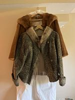 Front view of the two coats hanging on hangers, showing the brown faux fur coat behind the gray textured coat with fur collar.