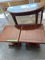 Photo showing one half moon-shaped console table behind two square wooden side tables on floor, all brown wood finish.