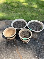 Four decorative garden pots on outdoor ground, three filled with soil, one smaller with matching saucer.