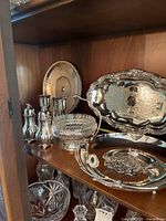 Assortment shown on wooden shelf includes various silver-plated trays, cut glass bowl, condiment set, water jug, and candle holders