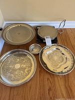 Four round silver plated serving trays of varying sizes and one silver plated trivet with handle placed on a wood floor against wall