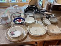 Overview of assorted frying pans, glass casserole dishes, metal roasting pan, bowls, teapot and plates on kitchen counter