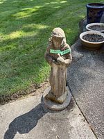 Full view of the statue standing on pavement beside grass with nearby flower pots, showing overall form and height.