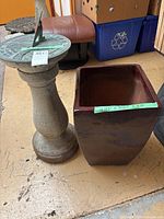 Full view of the concrete sundial next to the large brown ceramic planter. The sundial has a wide pedestal base and the planter is square-shaped with dark brown glaze.