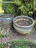 Two green garden pots placed outdoors by a wire fence surrounded by plants and flowers. One pot has a matching round tray underneath. Both show dirt and outdoor wear.