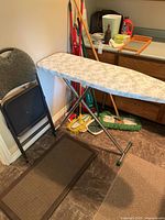 Photo of ironing board, two folding chairs, and mop and broom cleaning tools against wall on floor.