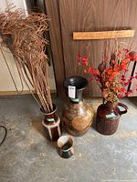 Photo showing four decorative ceramic vases on a floor near a wall, three are large sizes with various dried branches and flowers inside, the fourth is smaller and empty.