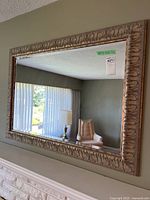 Large rectangular mirror with ornate carved frame hanging on a wall, reflecting an armchair, lamp, and window.