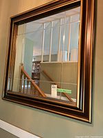 Beveled square wall mirror hung on a wall showing reflection of a staircase and light fixture.