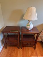 Two wooden nightstands side by side with one holding a cream colored lamp with pleated shade on top, sitting on hardwood floor against a beige wall.
