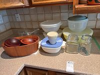 Overview of all items in the lot arranged on kitchen counter showing wood, glass, and plastic bowls, storage containers, jars with lids, and utensils.