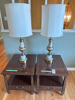 Two matching wooden side tables with single drawers, dark wood finish, topped with two gold and cream colored lamps with white cylindrical shades.