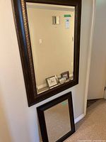 Photo showing a large rectangular mirror with an ornate dark brown frame hanging on the wall, and a smaller square mirror below it on the floor, also framed.