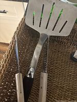 Photo showing a slotted spatula, grilling fork, and knife with light wood handles placed together on a woven surface.