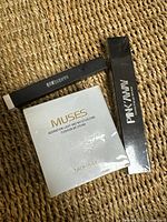 Top view of the closed boxes arranged on a woven surface showing the front face of the BB cream and mascara packaging alongside the eyebrow pencil box.