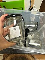 Photo showing a hand holding one clear glass jar with black lid and price label showing $1.99, inside a clear plastic box containing multiple similar jars.