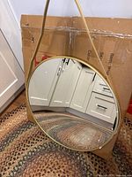 Round mirror with gold frame hanging by a strap, placed against a cardboard box background on a woven rug.