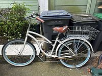 Full side view of white cruiser bike against backdrop of trash bins, showing frame, tires, seat, and pedals.