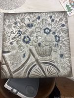 Full view of square metal wall decor depicting embossed floral basket on bicycle, silver with blue accents