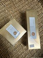 Two boxed Gold Beauty skin care products on woven textured surface. Left is Essence Cream, right is Essence Lotion, both in gold packaging with front labels visible.