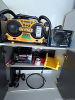 Cabinet shelf showing DeWalt radio-charger, OdorFree unit, MotoMaster charger, yellow tray, black rods and cables