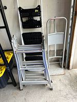 Overall view of three items: articulating ladder folded, Cosco step ladder behind, metal hand truck to right