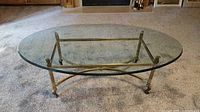 Oval glass coffee table shown on carpet with front view of brass base and clear glass top.