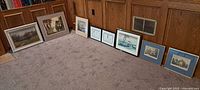 Nineteen framed wall art prints arranged on carpeted floor against wood-paneled wall showing a variety of art themes and frame styles; some frames appear wooden, others metal.